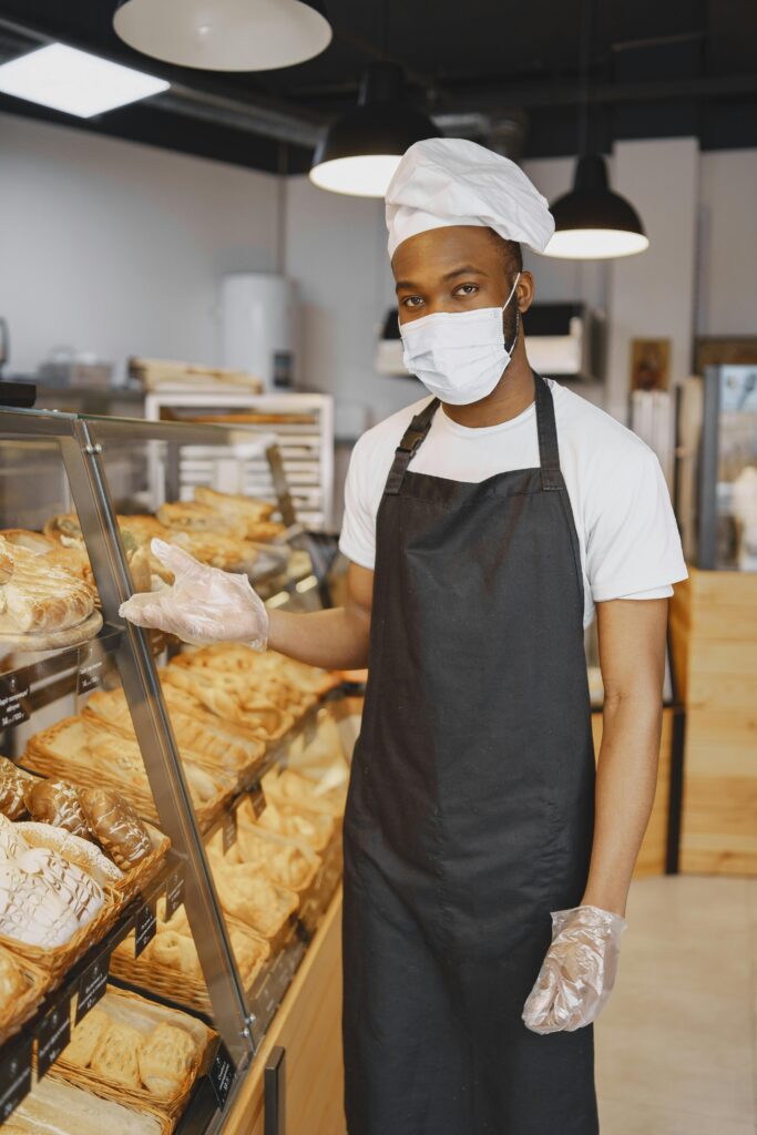 Masked baker presenting an assortment of fresh pastries in a bakery. Indoor shot.